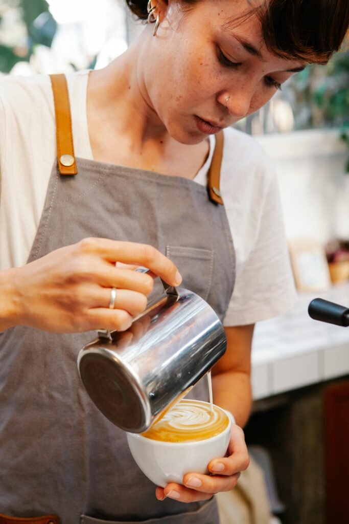 Primer plano de una barista con delantal gris vertiendo leche texturizada en una taza blanca para hacer un dibujo de café con leche.