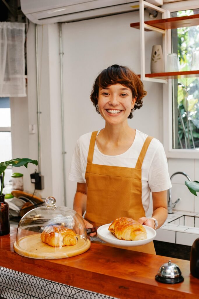 Lucía sonriendo en la barra de la cafetería con delantal mostaza mientras sirve un croissant en un plato blanco.