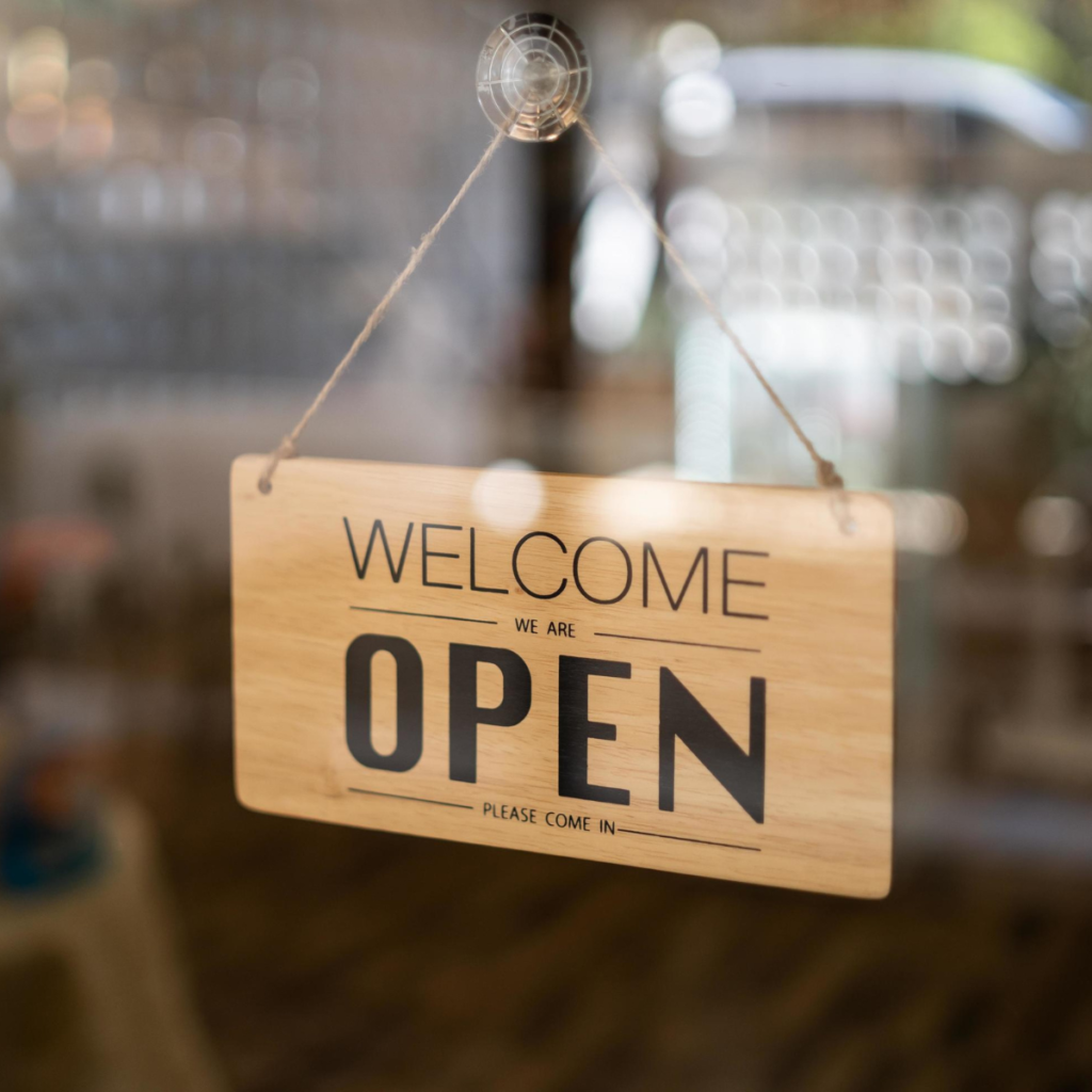 rimer plano de un letrero rectangular de madera colgado con una cuerda fina detrás del cristal de una ventana o puerta. El letrero tiene grabada la palabra "WELCOME" en la parte superior, seguida por la frase "WE ARE OPEN" en letras negras grandes y llamativas en el centro, y "PLEASE COME IN" en la parte inferior. El fondo del establecimiento aparece desenfocado.