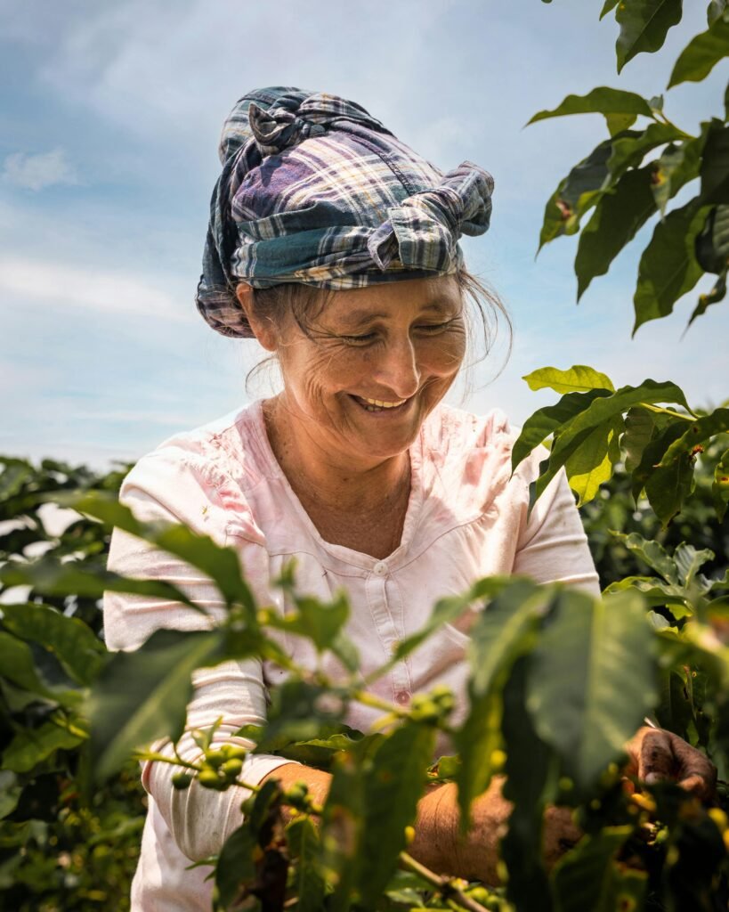 Retrato de una mujer sonriente con pañuelo a cuadros en la cabeza y camisa rosa, trabajando entre plantas de café con el cielo de fondo.