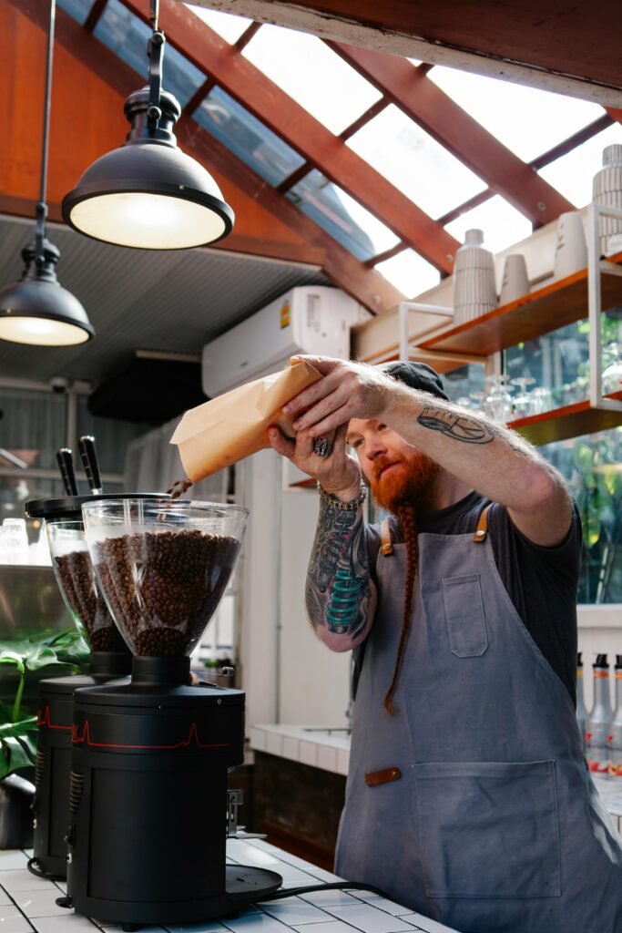 Primer plano de un barista con delantal gris echando granos de café tostado en la tolva de un molino profesional.