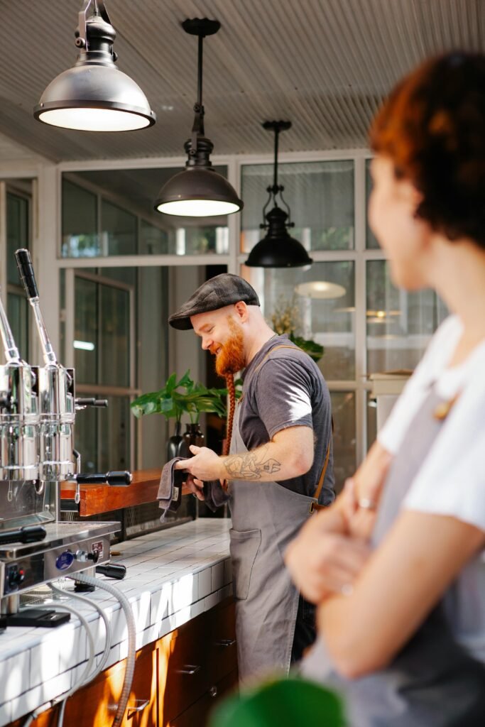 El barista Andrés sonriendo y limpiando la barra de café con un paño, visto desde la perspectiva de su compañera en primer plano.