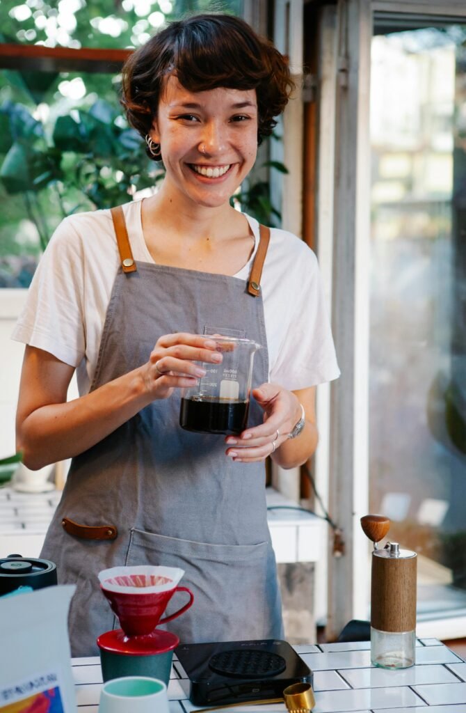 Retrato de la barista Lucía sonriendo en la barra mientras sujeta un vaso de cristal con café de especialidad recién preparado.