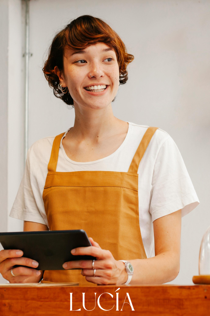 Retrato sonriente de la barista Lucía con delantal mostaza sosteniendo una tablet en el mostrador del local.